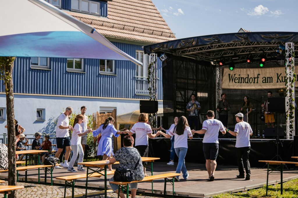 Um Gelder für das Gemeindehaus für alle zu finden, könnten Verantstaltungen organsiert werden. Foto von <a href="https://unsplash.com/de/@theodortietze?utm_source=unsplash&utm_medium=referral&utm_content=creditCopyText">Theodor Tietze</a> auf <a href="https://unsplash.com/de/fotos/menschen-die-bei-einem-festival-im-freien-im-kreis-tanzen-5DgkfM4M2K0?utm_source=unsplash&utm_medium=referral&utm_content=creditCopyText">Unsplash</a>
